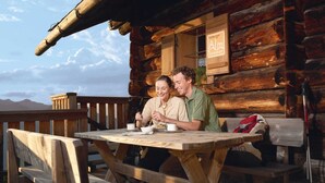 A wooden cabin with a rustic exterior, a wooden table with two people dining, and a mountain view in the background. - Large Apartment in Sankt Georgen With Balcony (Bruck an der Grossglocknerstrasse)