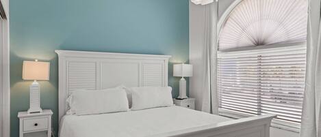A bedroom with a white bed, two bedside tables with lamps, a ceiling fan, and a window with blinds.