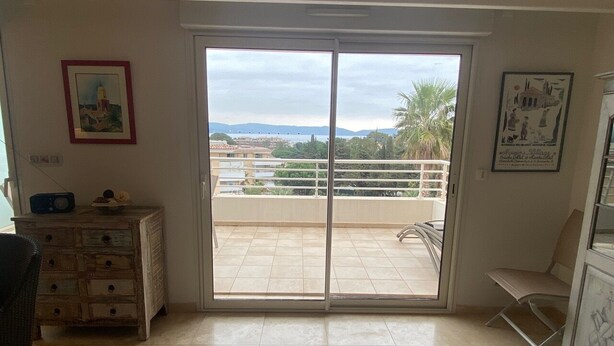 A room with a sliding glass door leading to a balcony with a view of palm trees and buildings. There is a wooden dresser with drawers and a chair. - 5-minute walk to beaches, shops, sea view, air-conditioning, pool, 79 m2 T3, garage. (Cavalaire-sur-Mer)