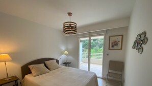 A bedroom with a large bed, a bedside table with a lamp, a chair, and a view of the outdoors through a sliding door. - 5-minute walk to beaches, shops, sea view, air-conditioning, pool, 79 m2 T3, garage. (Cavalaire-sur-Mer)