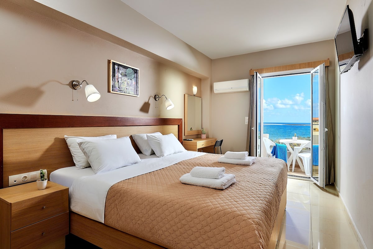 Una habitación de hotel con cama, mesita de noche, lámpara y vista al mar a través de una puerta abierta.