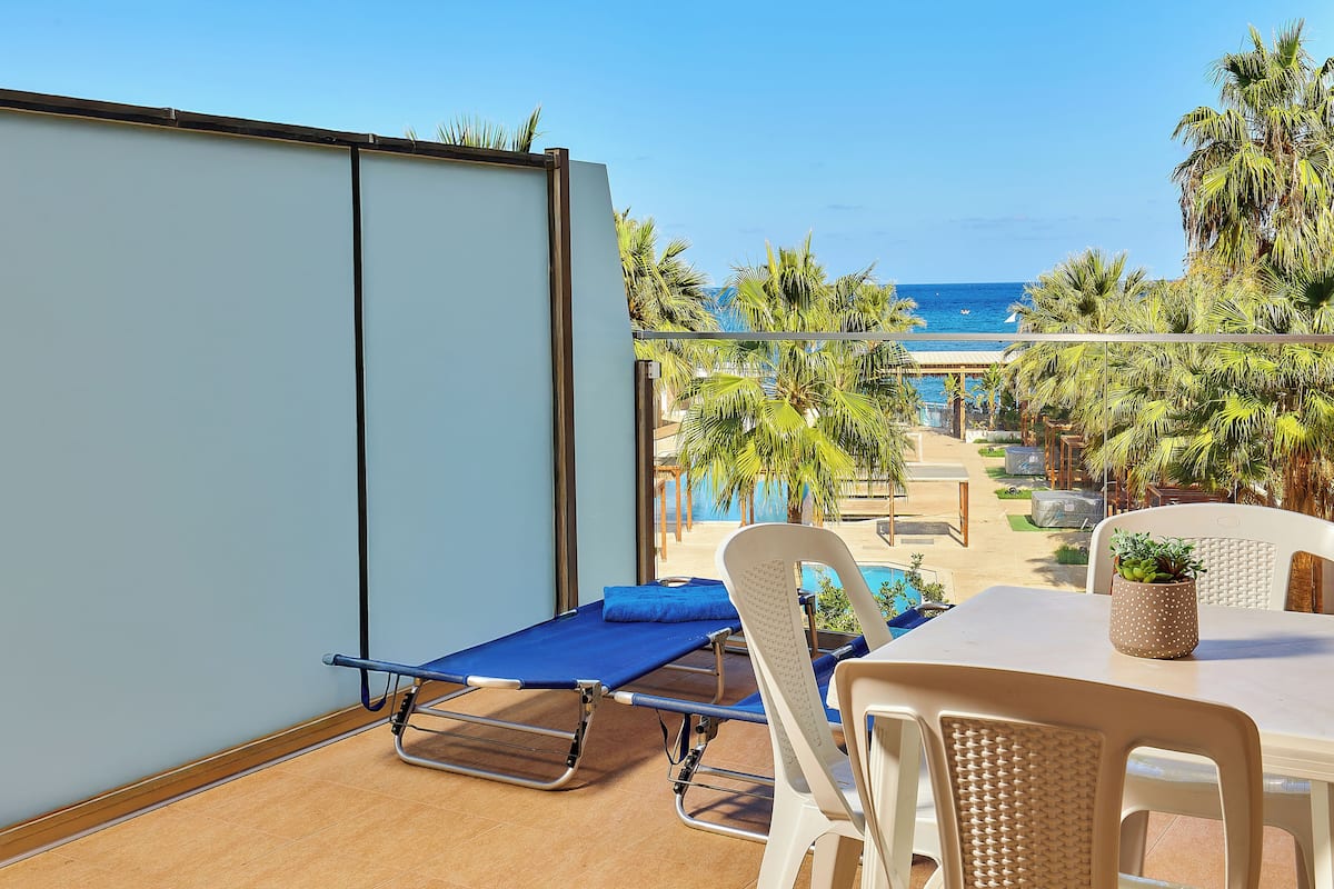 Un balcón con una mesa de masaje azul, sillas blancas y vistas a la playa y palmeras.
