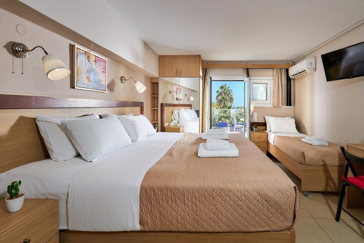 Une chambre d’hôtel avec un grand lit, un plus petit lit, une télévision, un balcon avec vue et un ventilateur de plafond.