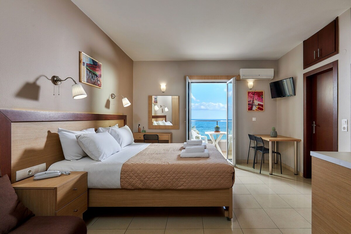 Une chambre d’hôtel comprenant un lit, une table de chevet, une lampe, une œuvre d’art, un bureau avec une chaise et une vue sur la mer.