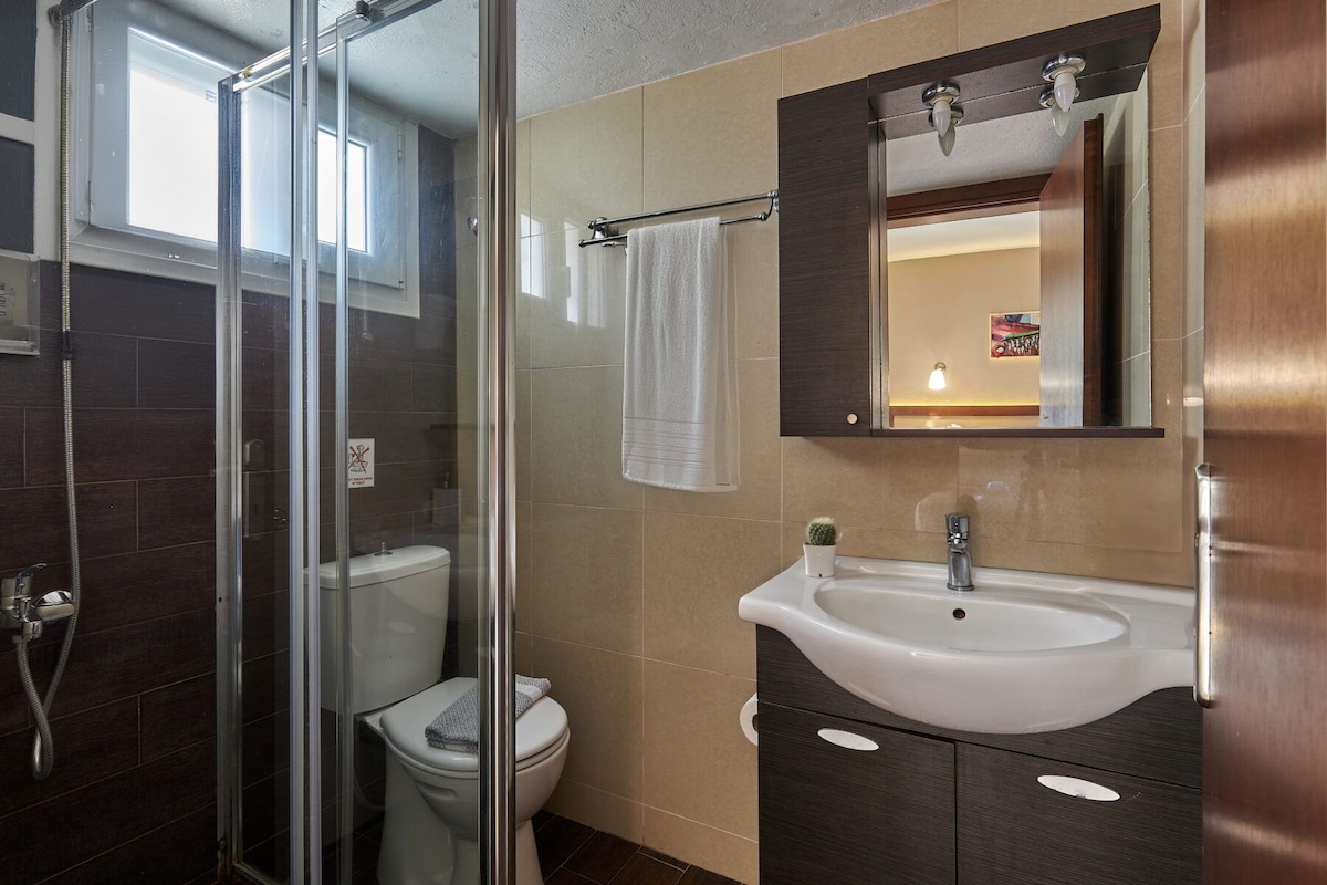 A bathroom with a glass shower enclosure, a toilet, a sink with a mirror above it, and a small potted plant.