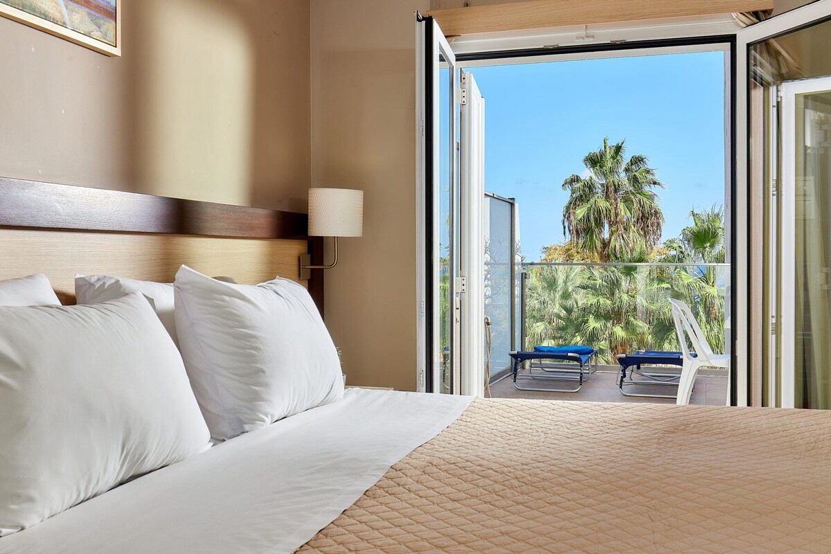 Une chambre d’hôtel avec un lit, une table de chevet, une lampe et un balcon donnant sur des palmiers.