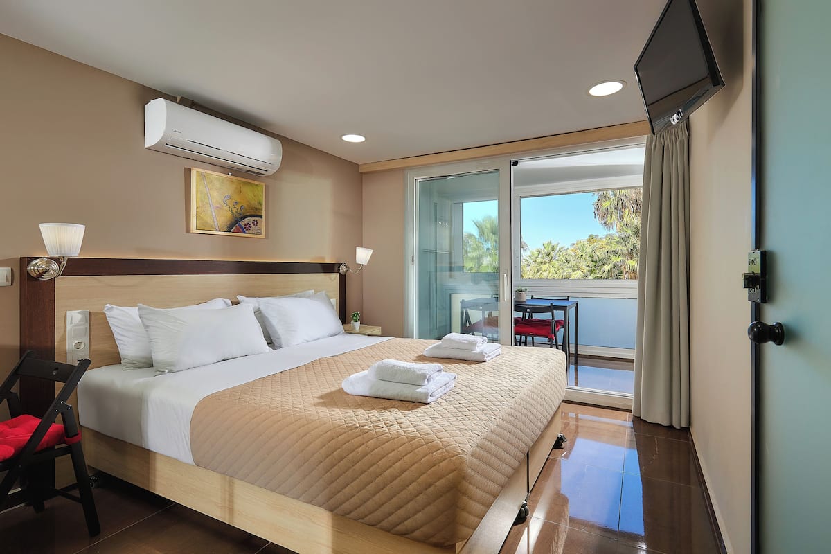 Habitación de hotel con cama, televisión, aire acondicionado, un cuadro y balcón con vistas.
