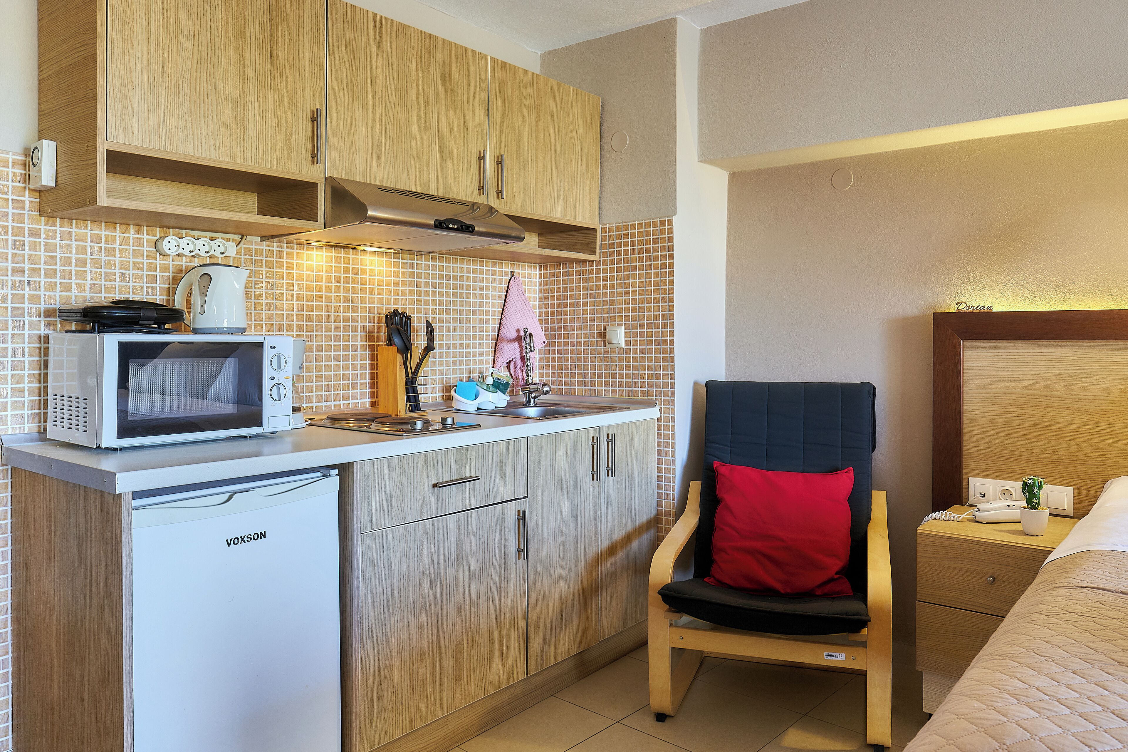 Una kitchenette compacta con microondas, tetera y tostadora, un armario de madera y una silla con un cojín rojo.