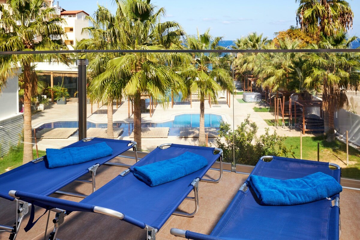 A balcony with three sun loungers, a pool, and palm trees.
