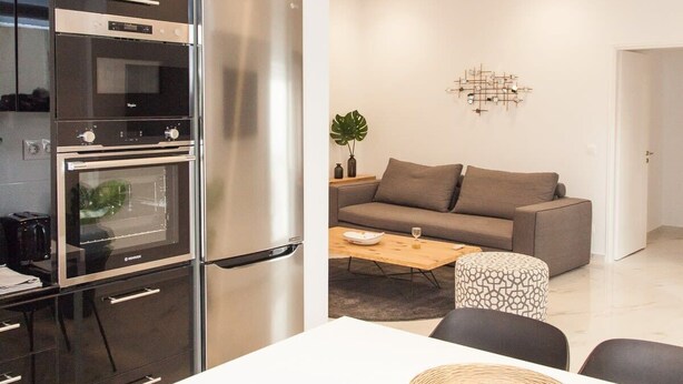 A modern kitchen with a built-in oven and refrigerator, a dining area with a white table and black chairs, and a living room with a gray sofa and a coffee table. - Comfortable Apartment in Athens Downtown (Athens)