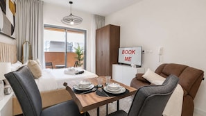 Ein Hotelzimmer mit Bett, Esstisch mit Tellern und Besteck, einer Couch, einem Fernseher und einem Schild mit der Aufschrift 'BOOK ME'.