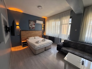 A modern hotel room with a bed, a sofa, a small table, and a window with curtains.