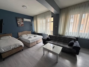 Quarto de hotel com duas camas, um sofá, uma mesa de centro e janelas amplas com cortinas.