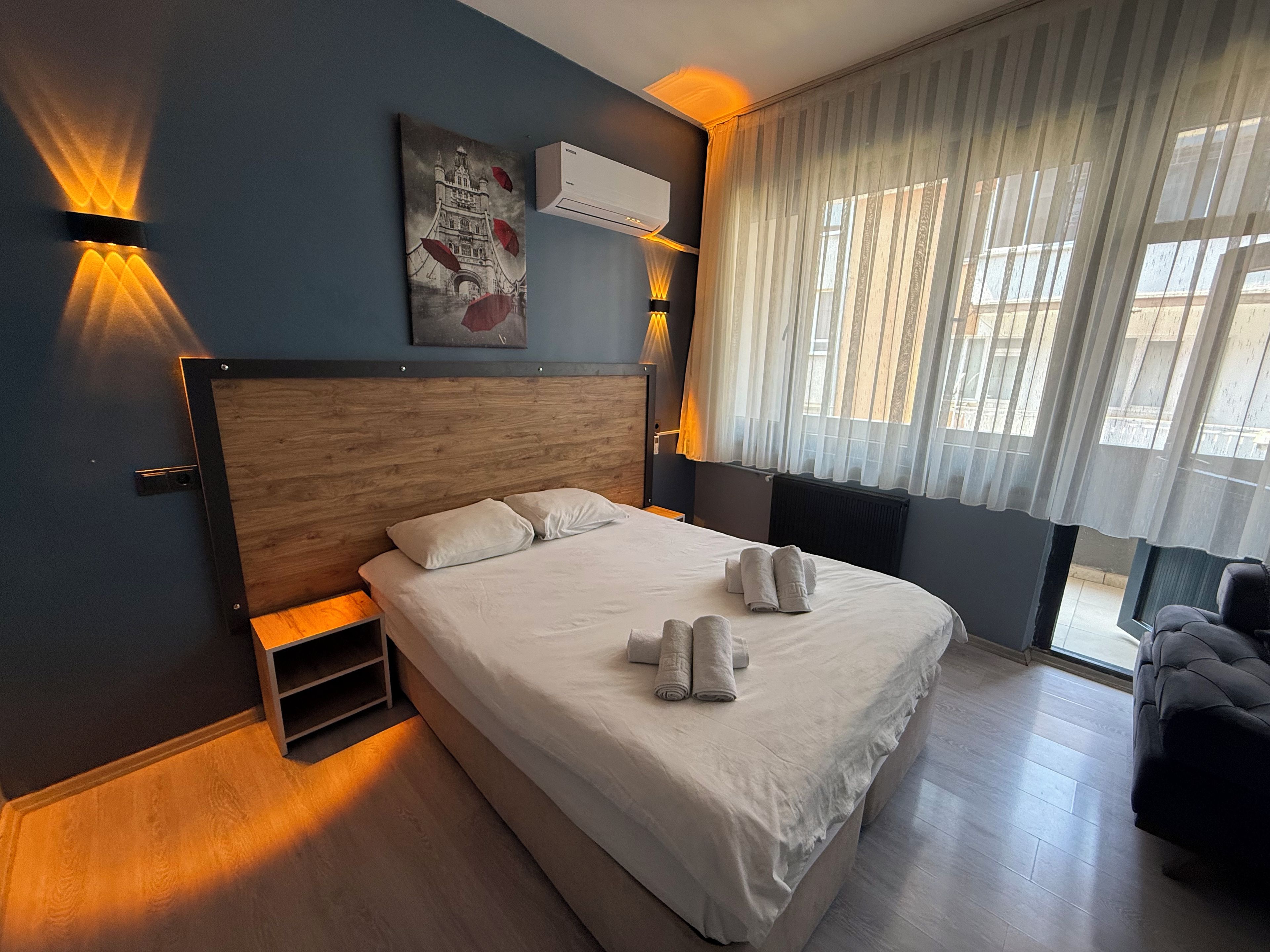 A hotel room with a large bed, a nightstand, a wall-mounted air conditioner, and a window with sheer curtains.