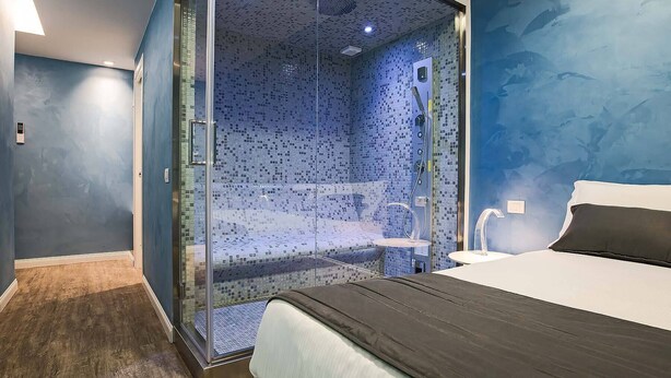 Una habitación de hotel moderna con ducha envuelta en vidrio, cama con cabecero oscuro y pared azul con textura.