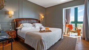 A spacious bedroom with a large bed, two bedside lamps, a wooden nightstand, a small table with a tray of breakfast, and a view of the outdoors through a large window. - Luxury Oceanfront at Reid's Gardens (Funchal)