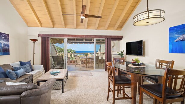 A spacious living area with a sliding glass door leading to a balcony, a dining table with chairs, a sofa, and a TV mounted on the wall.