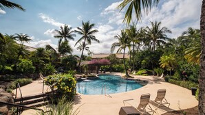 A pool surrounded by palm trees and greenery, with lounge chairs and a barbecue grill.