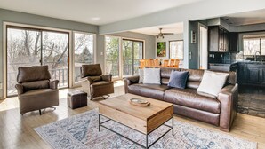 Un salón amplio con un sofá de cuero, sillones y una mesa de centro. Ventanas grandes permiten abundante luz natural y vistas al exterior.