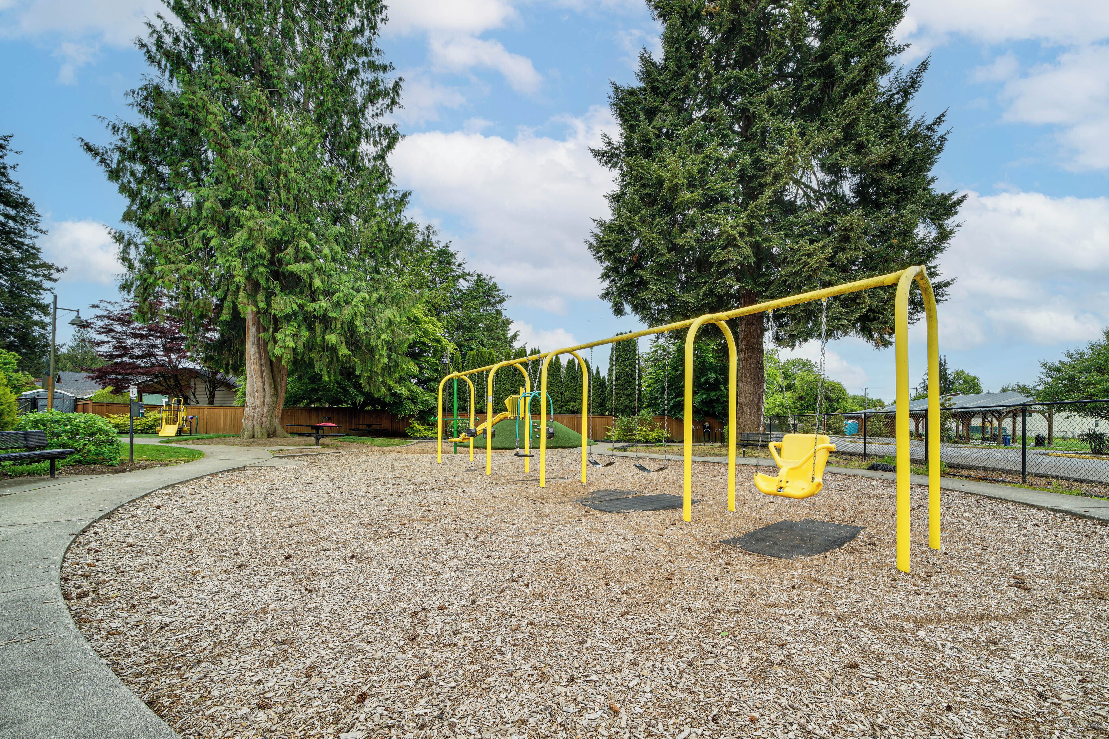 Un parque infantil con columpios y tobogán, rodeado de árboles y un camino pavimentado.