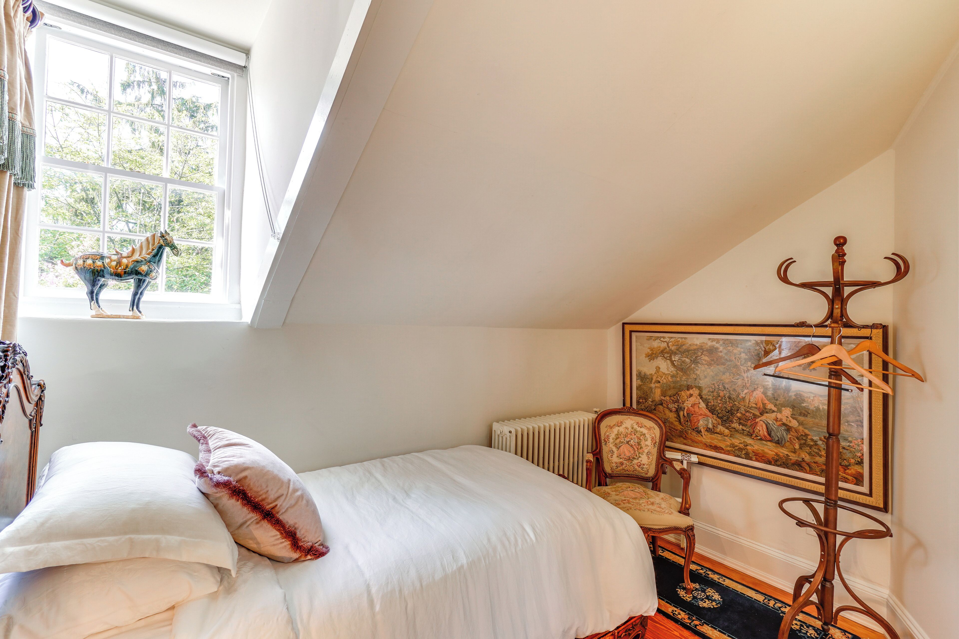 Un dormitorio con cama, radiador, silla, perchero de madera y ventana con vistas a árboles.