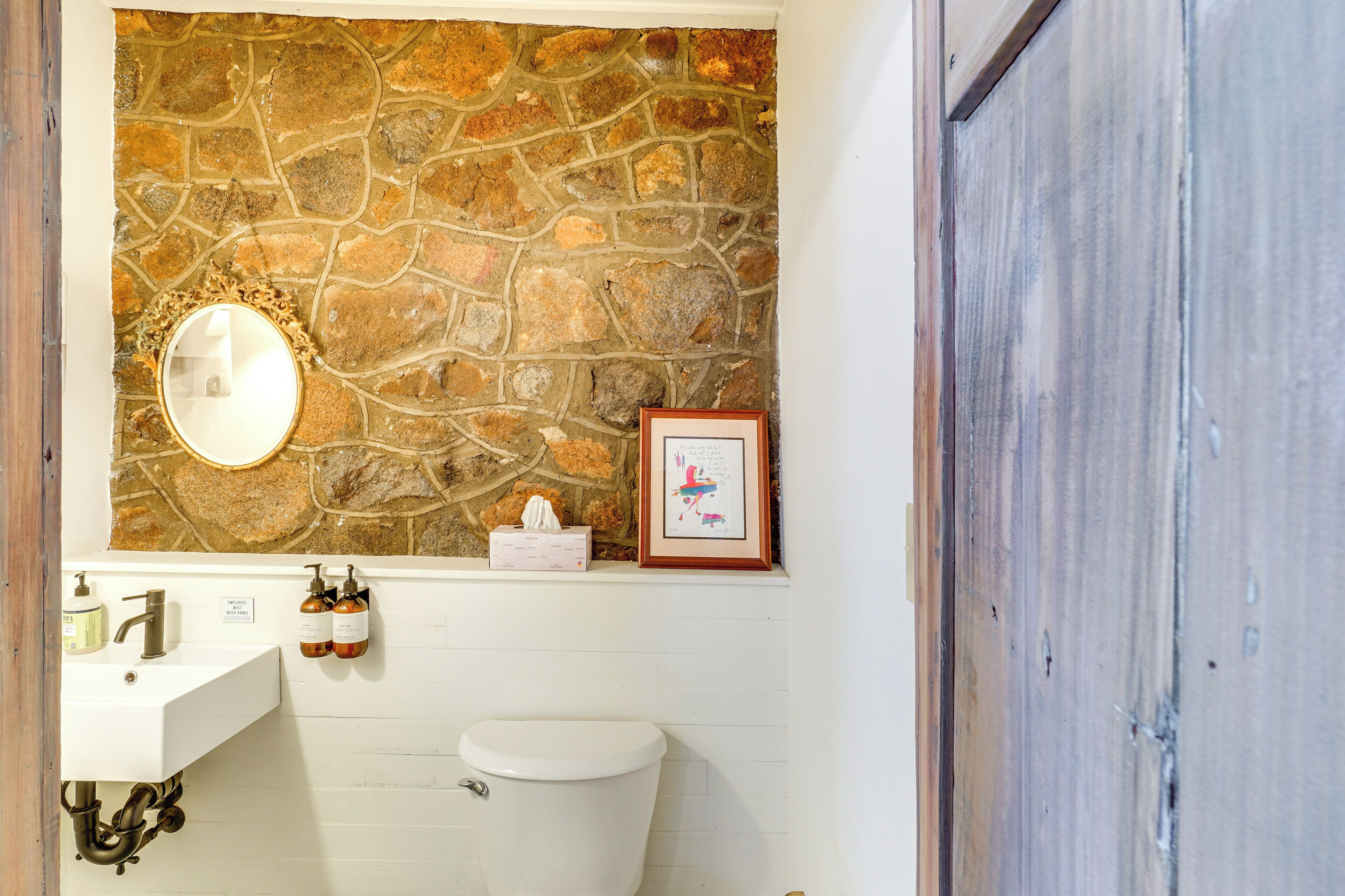 Ein Badezimmer mit Steinwand, Waschbecken in Weiß, einem Spiegel, einem gerahmten Bild und einer Toilette.