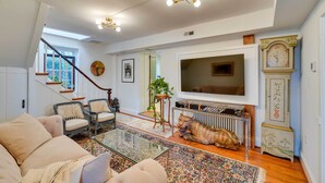 A living room with a sofa, chairs, a glass coffee table, a TV, a radiator, a clock, and a staircase. - Cottage w/ Furnished Patio & Garden: Dtwn Leesburg (Leesburg)
