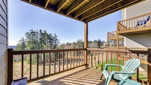 A wooden deck with two green Adirondack chairs, a wooden railing, and a view of trees and a building in the background. - Located in Snowshoe Resort: Townhome w/ Deck! (Snowshoe)