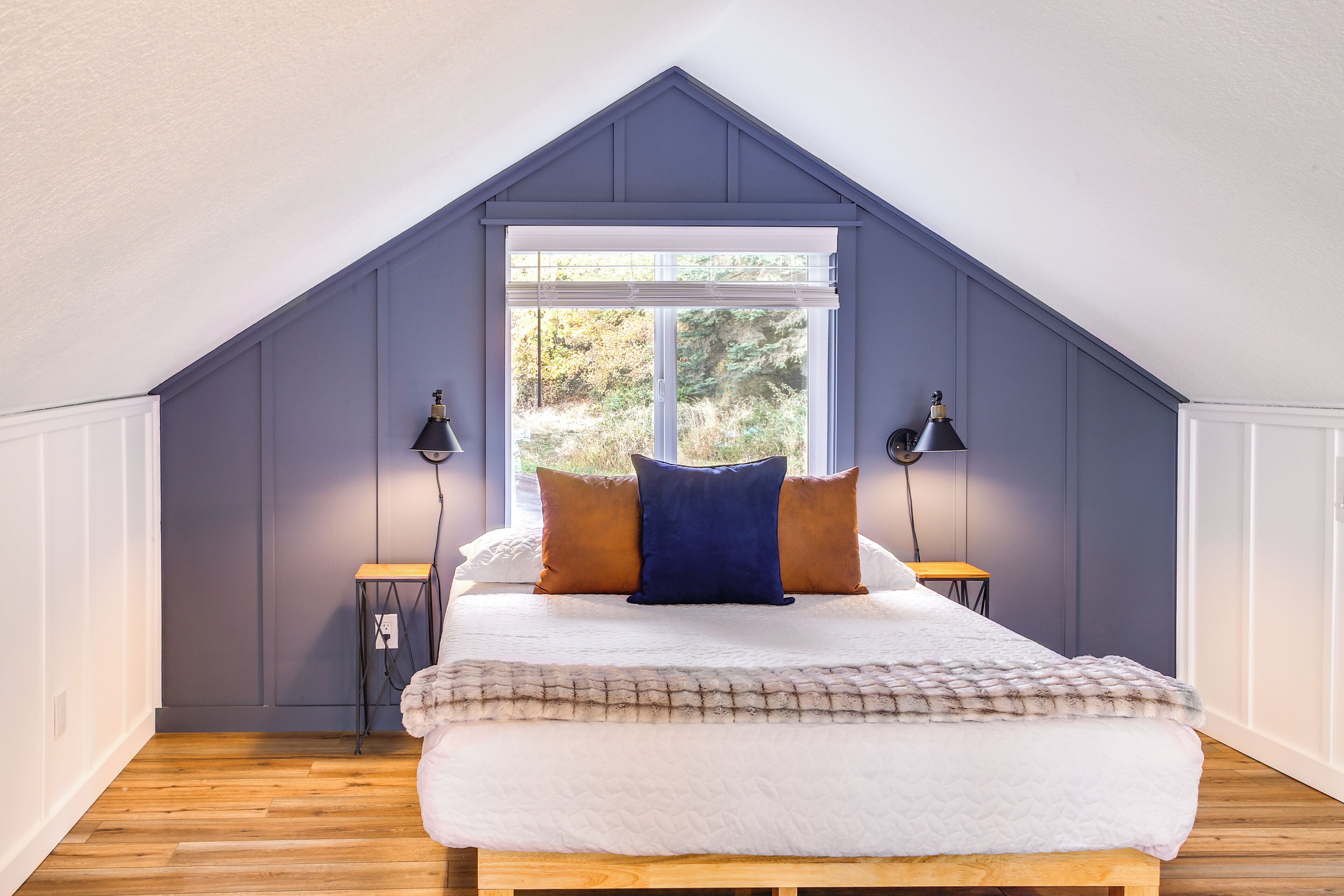 A bedroom with a sloped ceiling, a large window, a bed with white bedding, two bedside lamps, and a wooden floor.