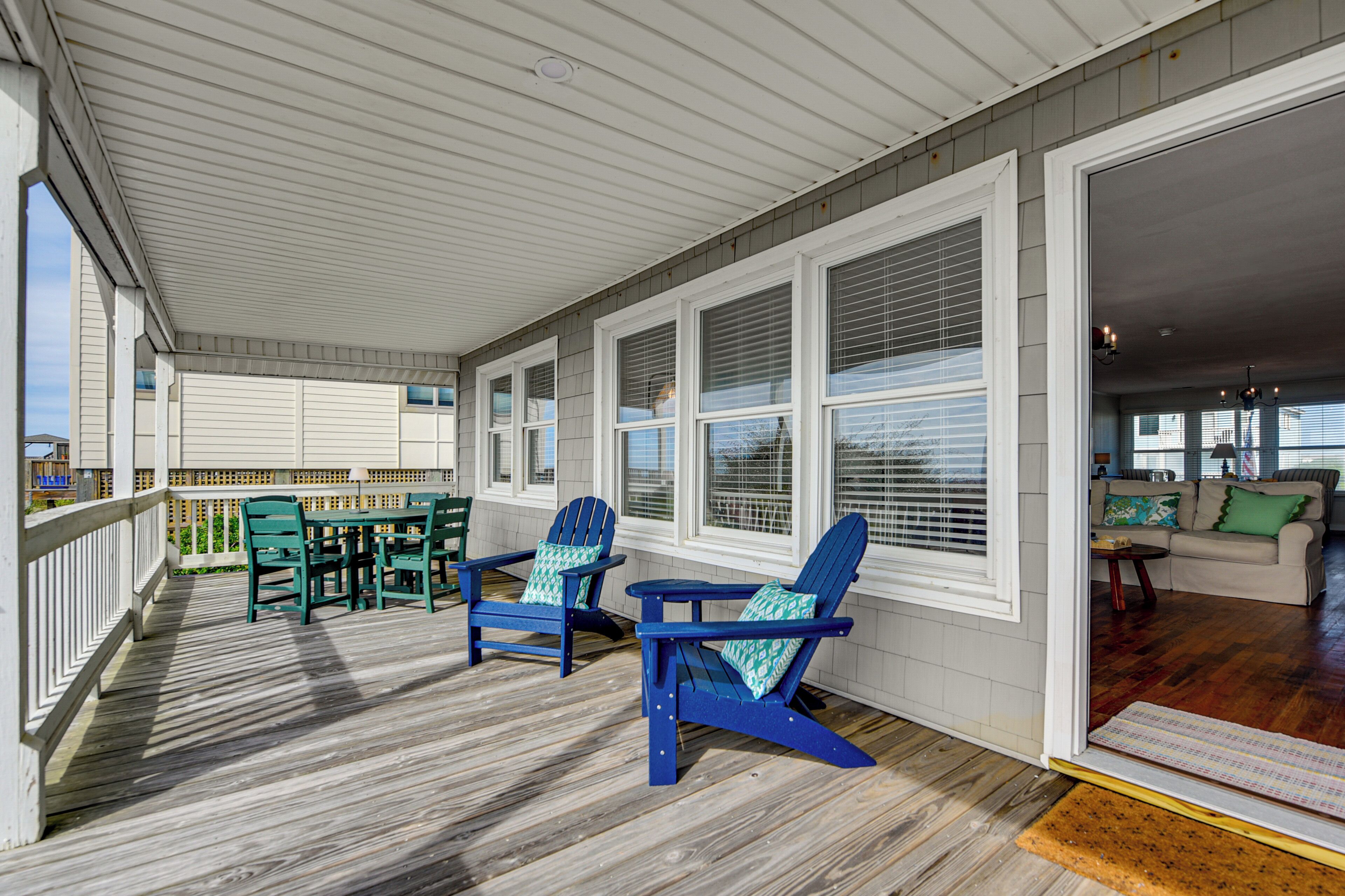 A covered porch with wooden flooring, two green Adirondack chairs, a table, and a view into the house.