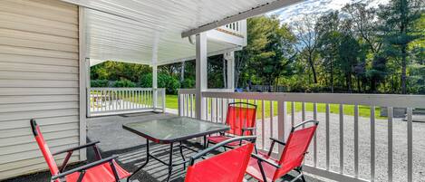 A covered outdoor patio with a table and red chairs, overlooking a grassy area with trees.