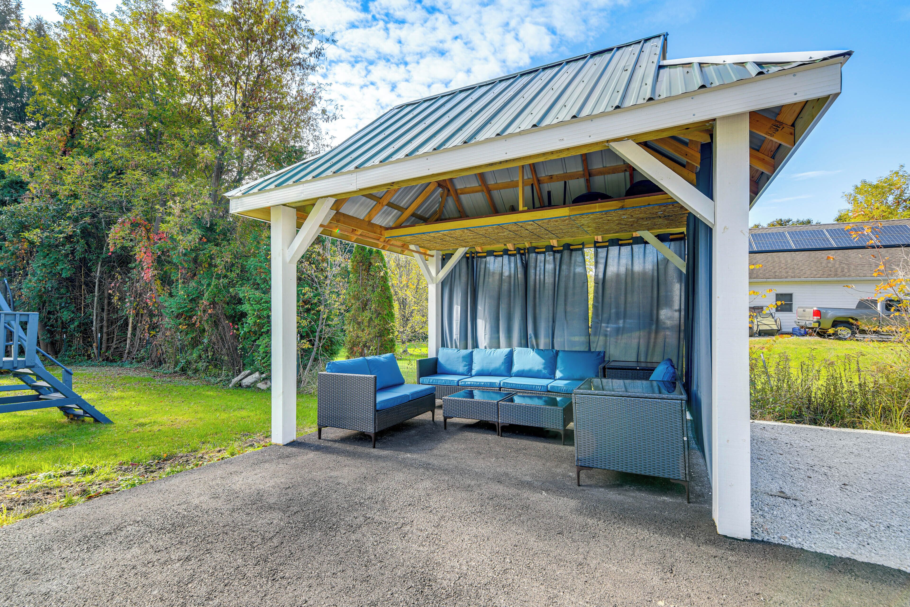Un espace extérieur aménagé, abrité et doté d’un canapé et de fauteuils, d’une structure en forme de pergola et d’une vue sur une maison équipée de panneaux solaires.