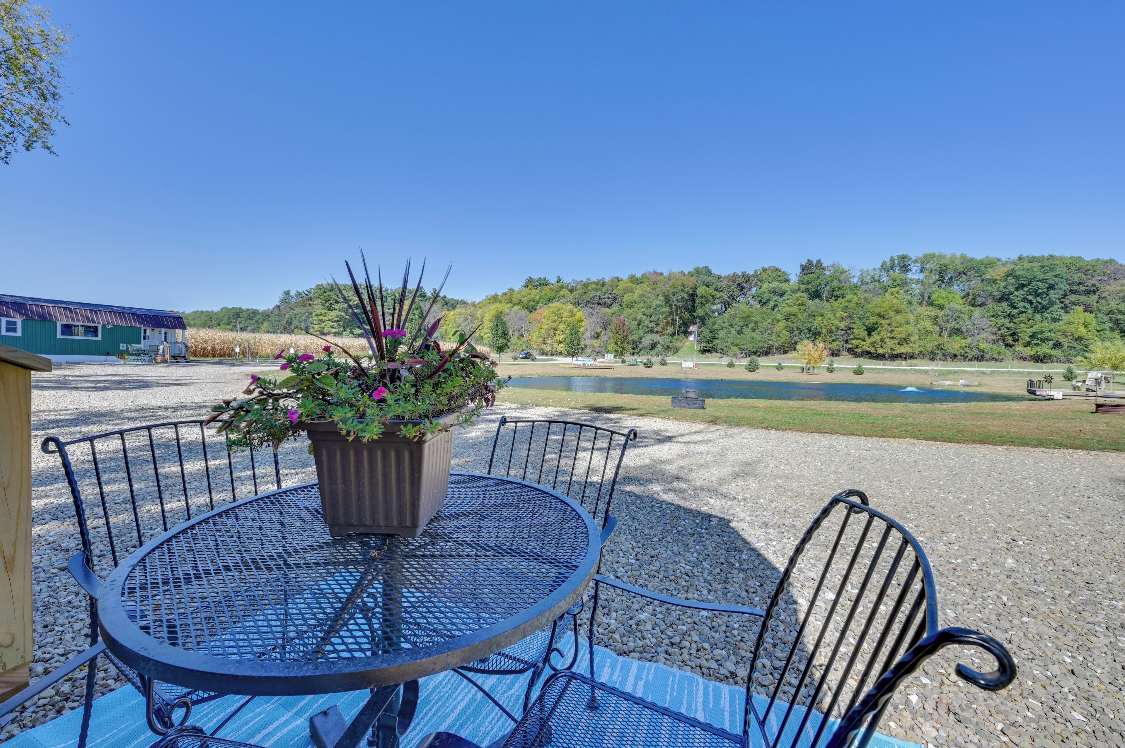 A round metal table with a potted plant in the center, surrounded by metal chairs, set on a gravel area with a view of a lake and trees in the background.