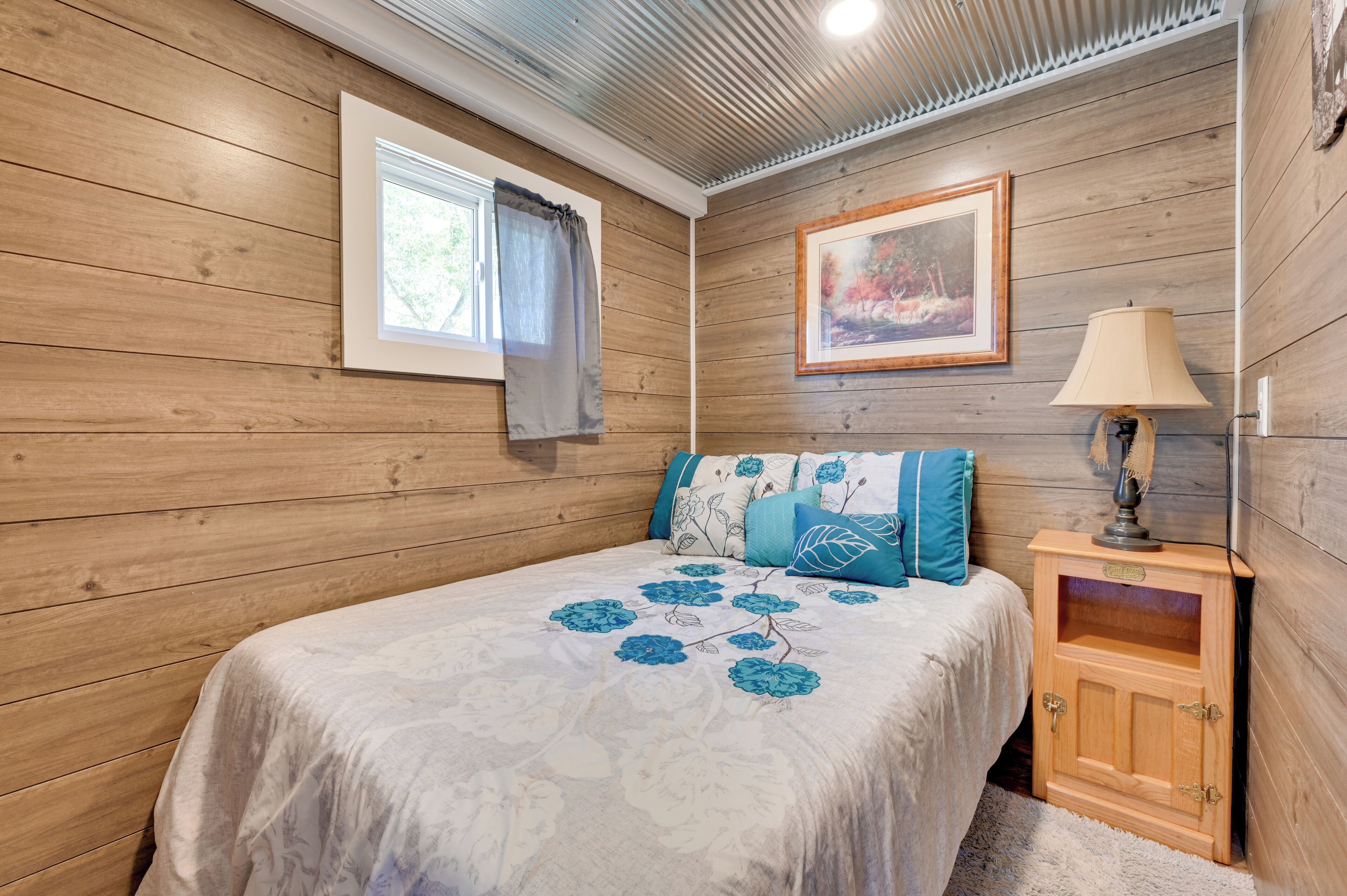 A bedroom with wooden walls, a bed with floral bedding, a nightstand with a lamp, and a window with curtains.