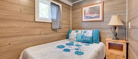 A bedroom with wooden walls, a bed with floral bedding, a nightstand with a lamp, and a window with curtains.