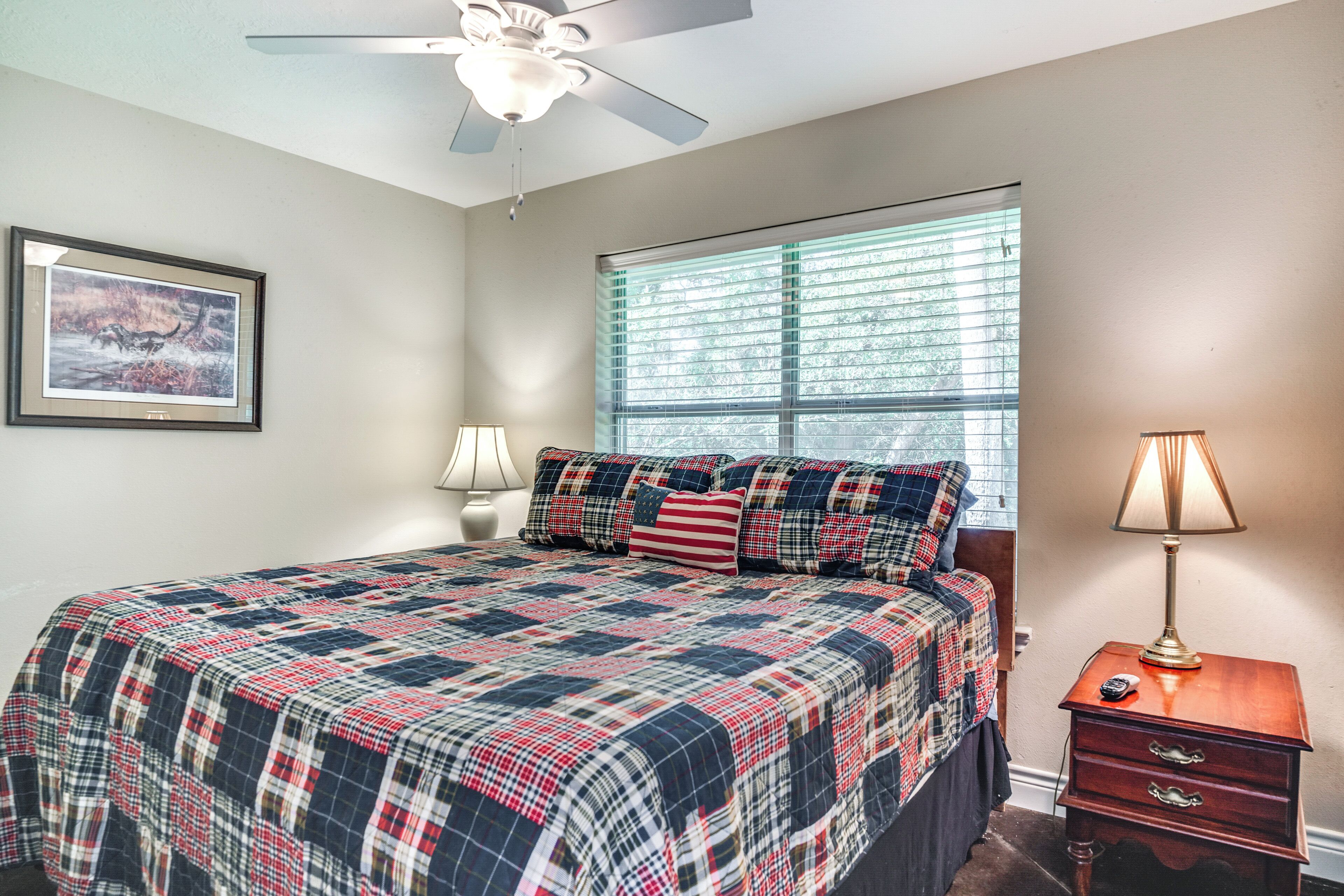 A bedroom with a bed, a ceiling fan, a window with blinds, a bedside lamp, and a framed picture on the wall.