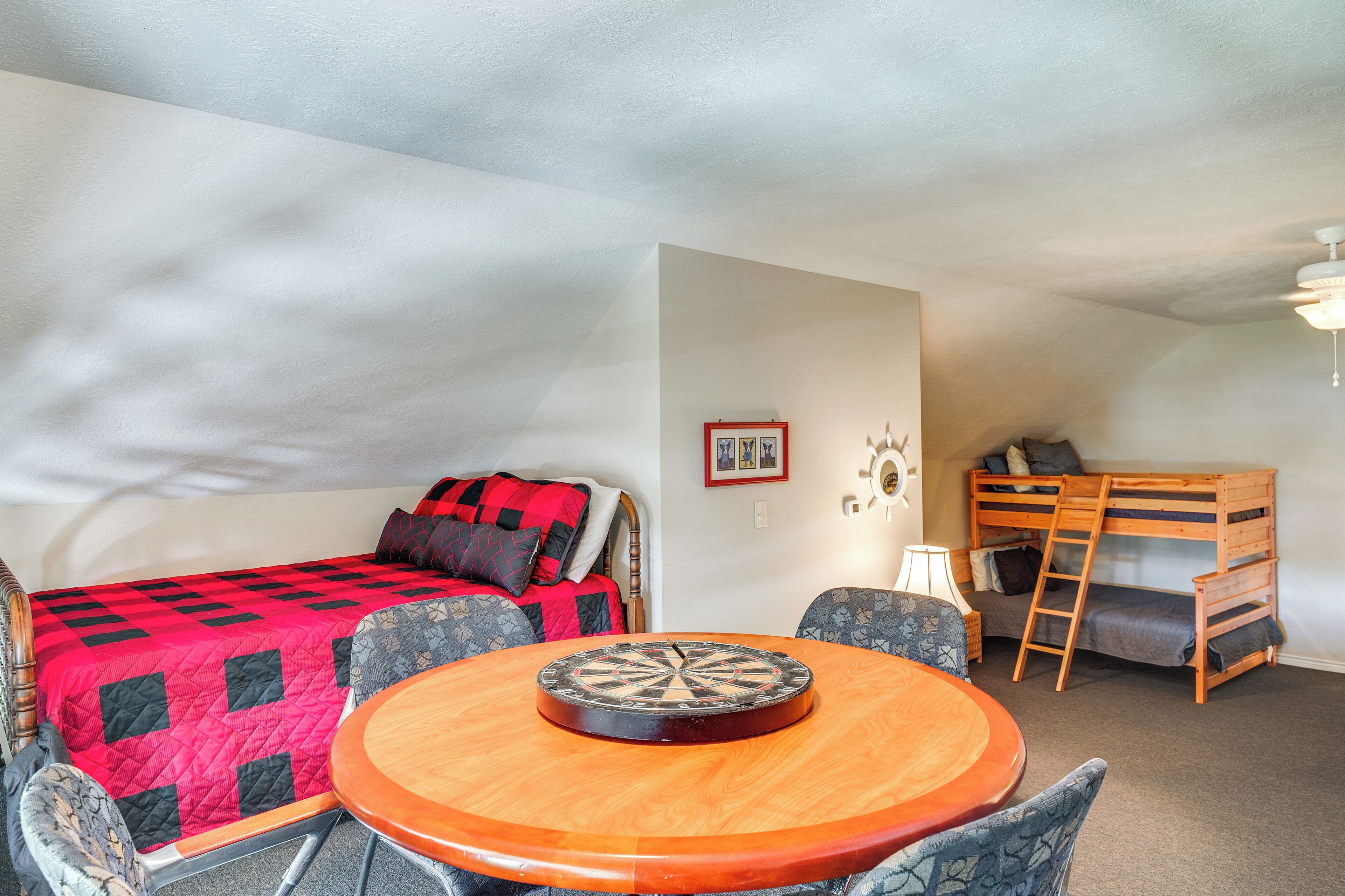 A room with a bed, a bunk bed, a round wooden table with a dartboard, and two patterned chairs.