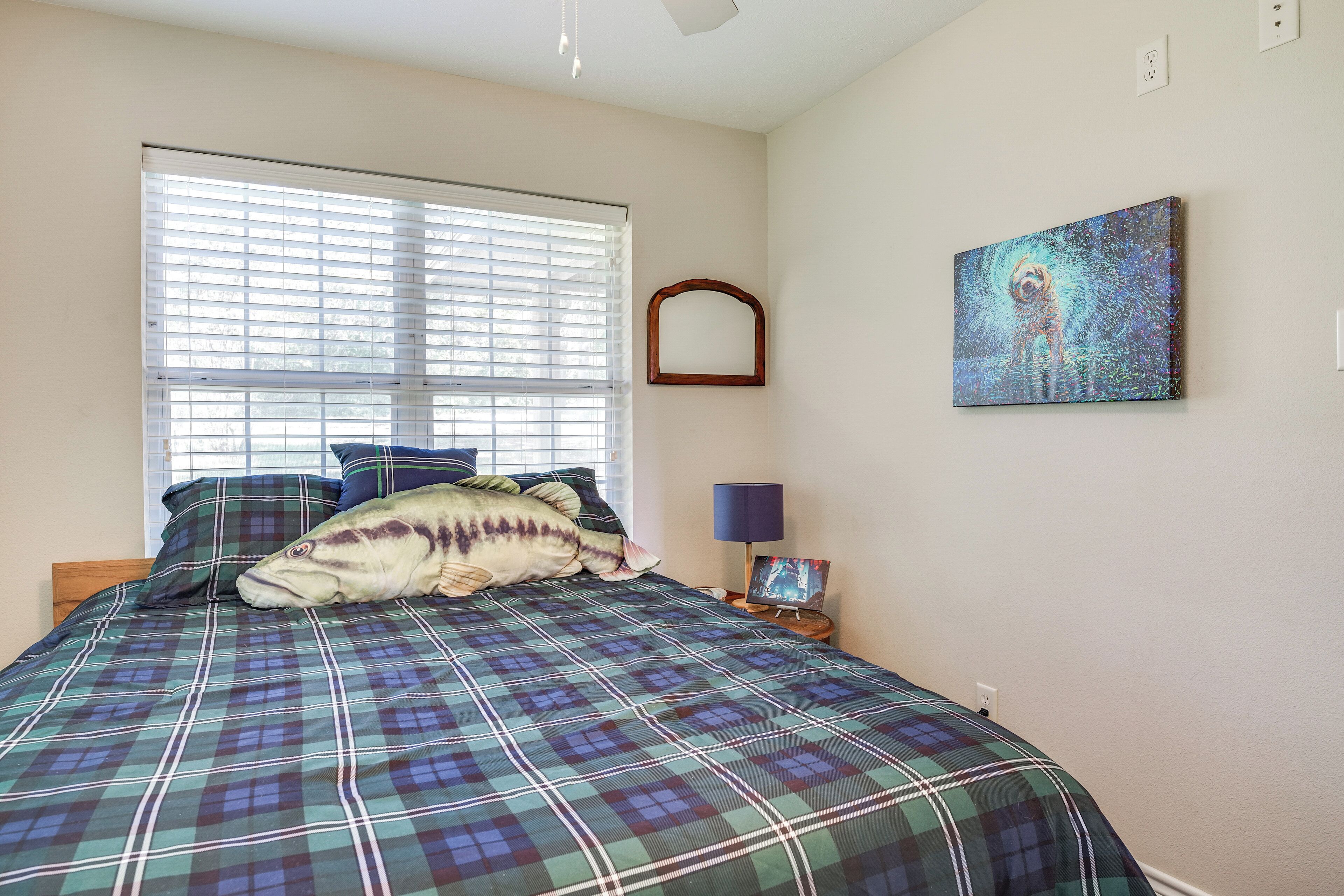 A bedroom with a plaid bedspread, a large window with blinds, a wall-mounted mirror, and a framed artwork.