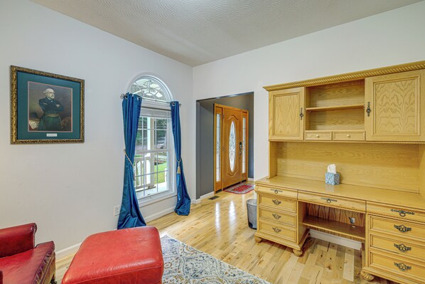 Una habitación con un escritorio de madera, una silla de cuero rojo, un retrato enmarcado y una ventana con cortinas azules.