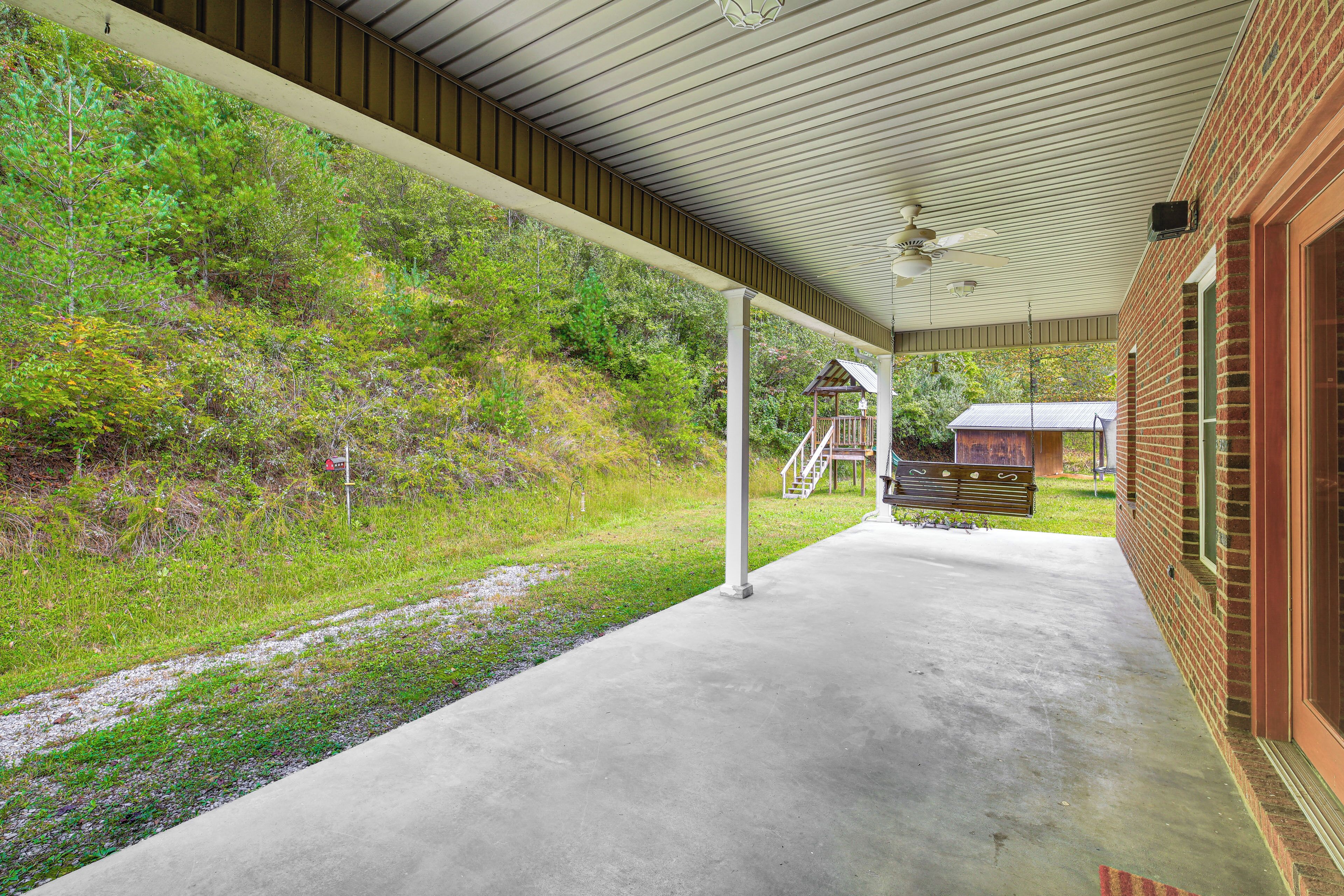 A covered outdoor area with a concrete floor, a brick building, and a wooden deck with stairs leading to a house.