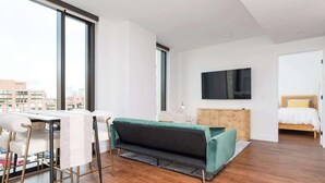 Quarto moderno e bem iluminado, com área de refeições, sofá, televisão na parede e cama com cabeceira de madeira.