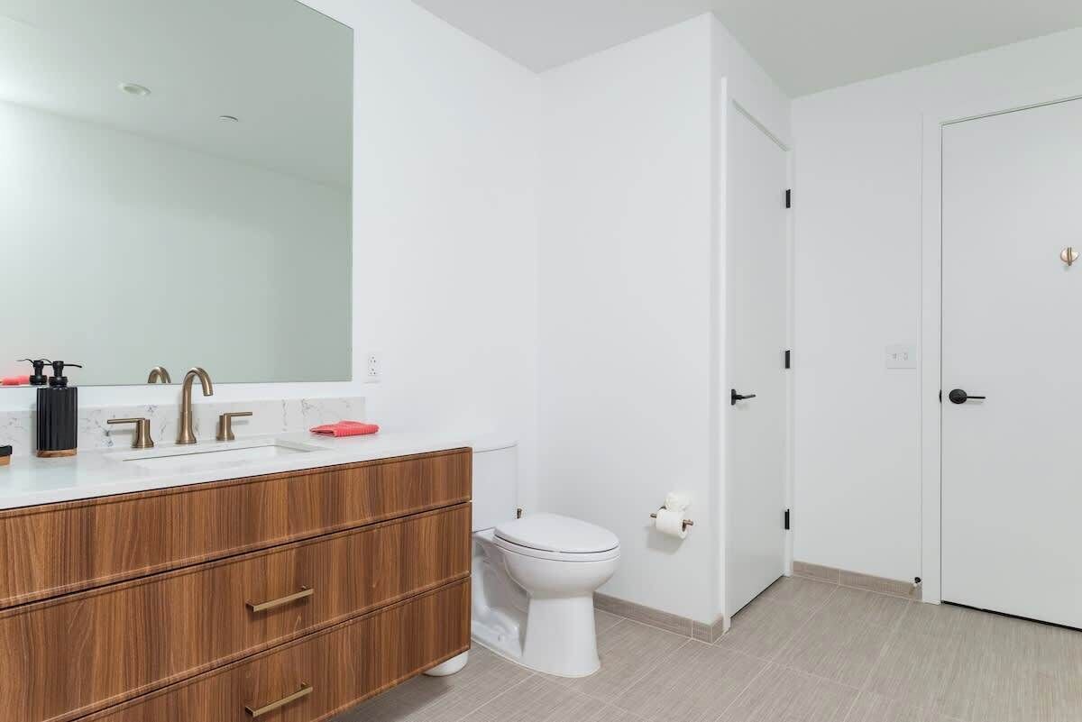 A modern bathroom with a white toilet, a wooden vanity with a large mirror, and a white sink.