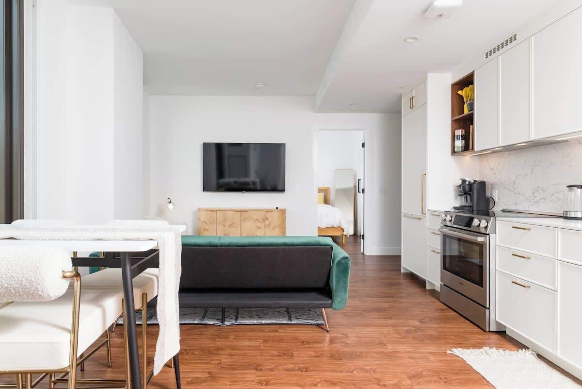 Et moderne, lyst værelse med spisebord, sofa, tv ophængt på væggen og et tekøkken med skabe og hårde hvidevarer.
