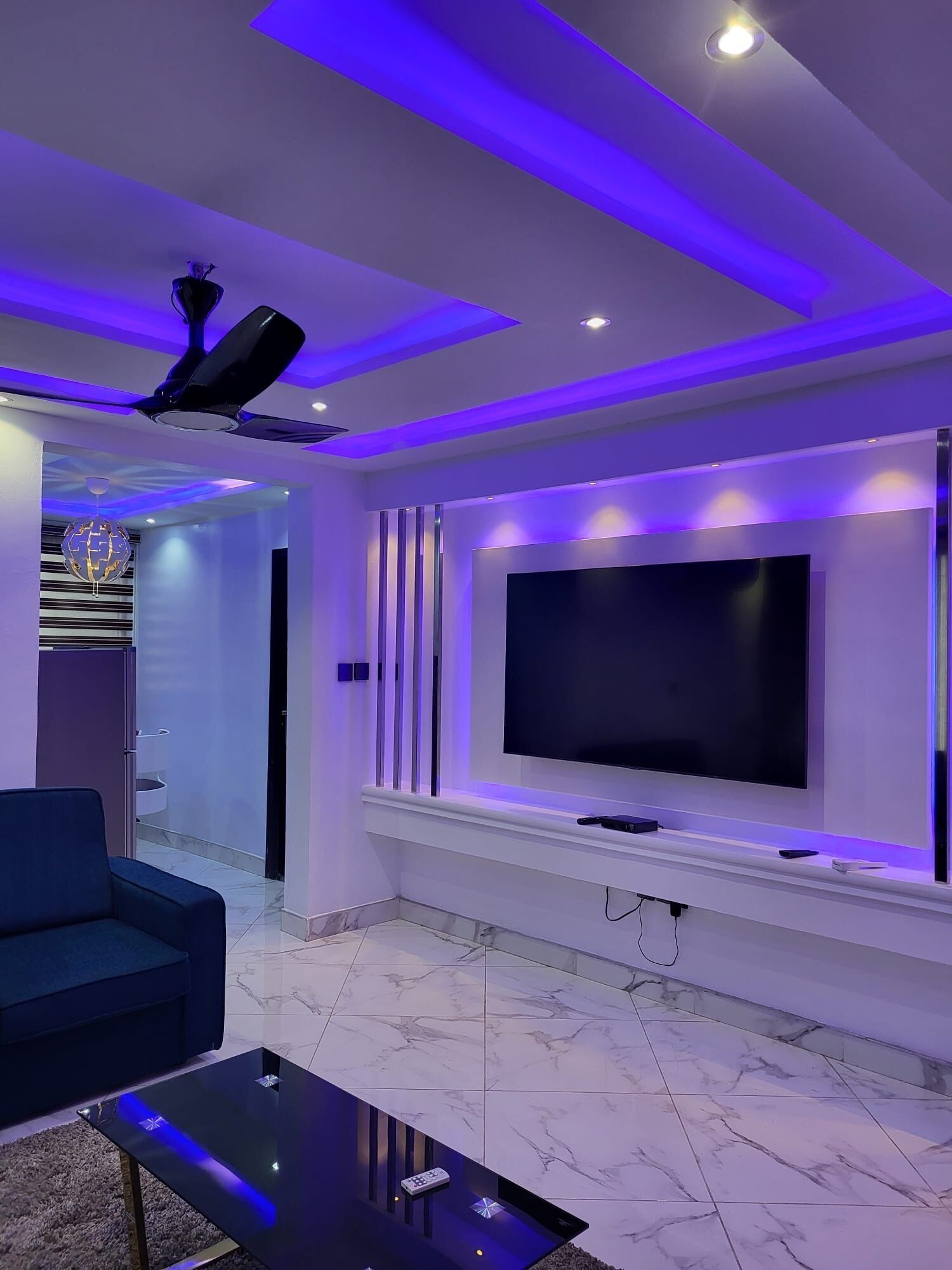 A modern living room with a large flat-screen TV mounted on the wall, a ceiling fan, and a marble-tiled floor.