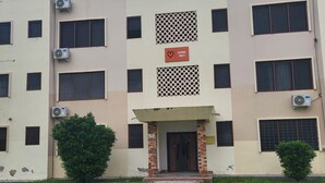 Un edificio de varios pisos con entrada central, unidades de aire acondicionado y un pequeño jardín.