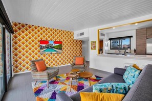 A modern living room with a patterned wall, a sofa, two armchairs, a coffee table, and a kitchen area with a refrigerator and sink.