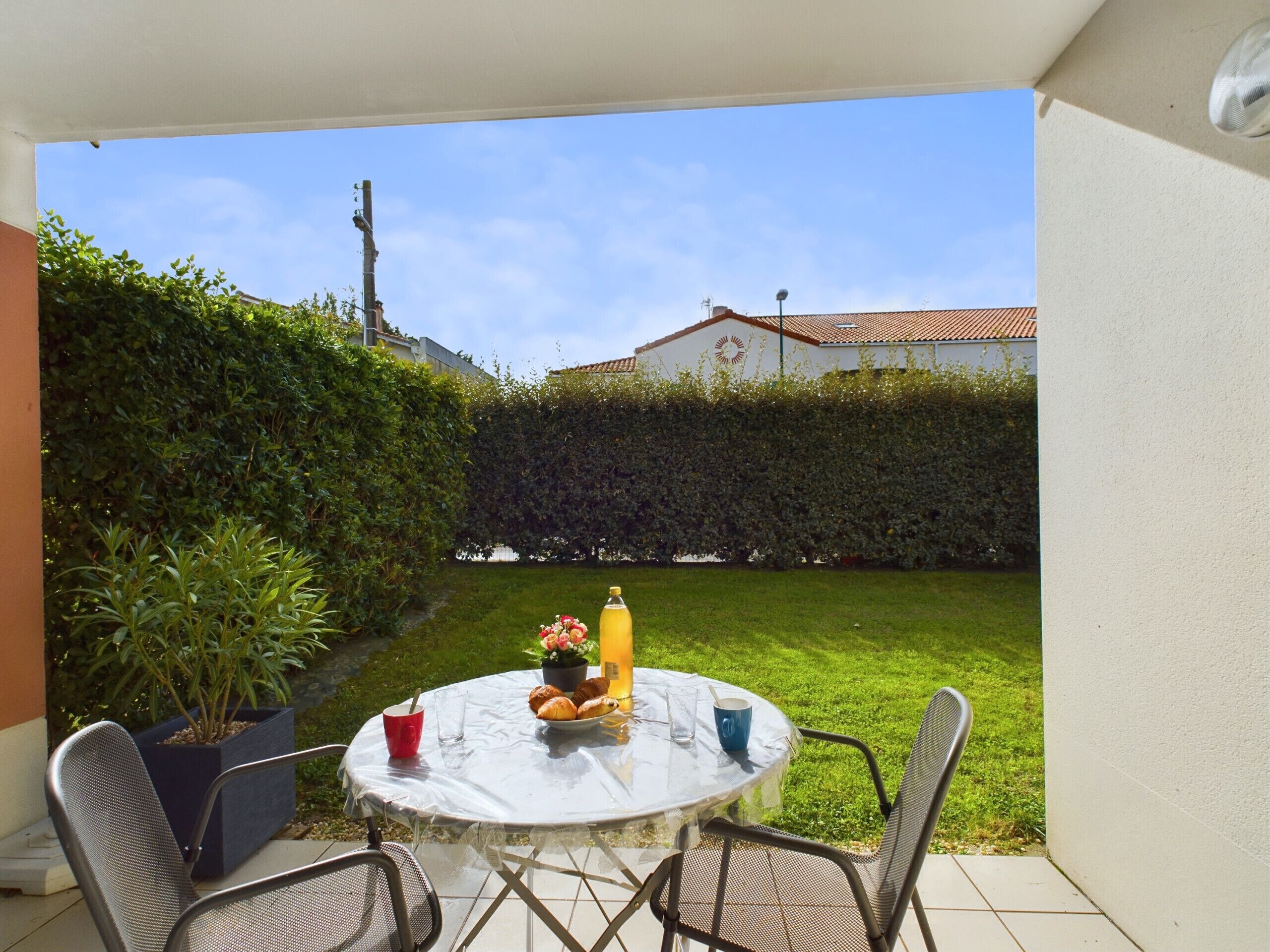Une table ronde recouverte d’une nappe blanche, deux chaises, une plante en pot, un verre de jus d’orange, une assiette de croissants et une tasse bleue.