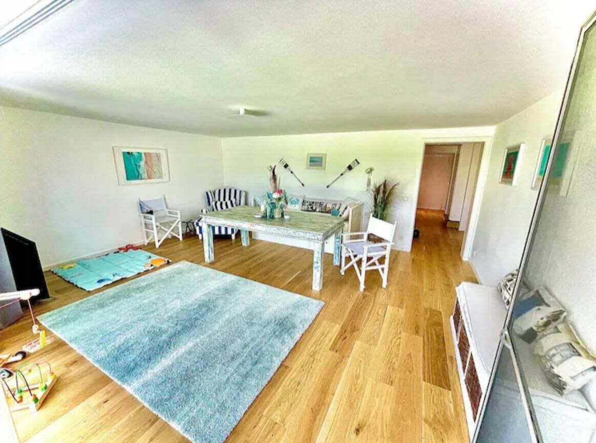 A spacious living room with a wooden floor, a dining table with chairs, a large rug, and a television.
