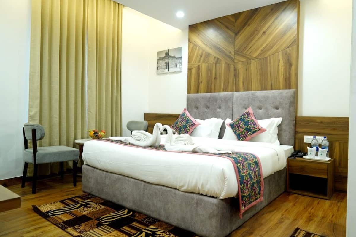 A hotel room with a large bed, a wooden headboard, a small table with a chair, a nightstand with a phone and water bottles, and a wall-mounted picture.