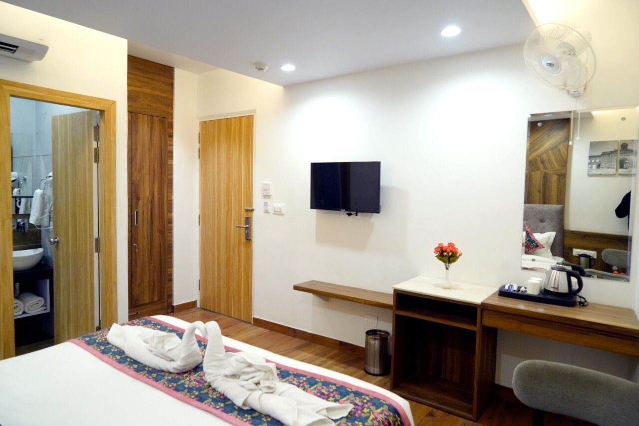 Habitación de hotel con cama, escritorio, televisión, espejo, estante pequeño y ventana.